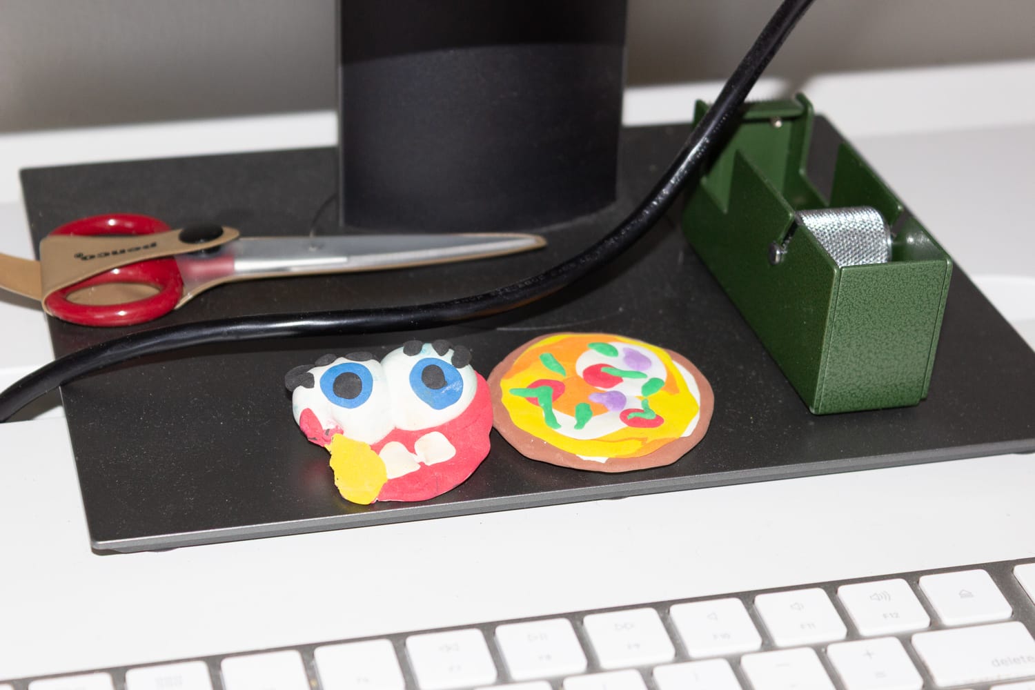 A tape dispenser, scissors, and two clay figurines under a computer monitor on a desk.